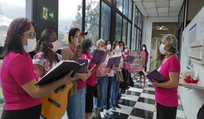 A foto mostra um grupo de mulheres perfiladas em um corredor envidraçado; elas usam máscara, vestem blusas cor-de-rosa, em sua maioria, e seguram livros abertos, exceto uma, que segura um violão; à sua frente está outra mulher, que conduz a ação do grupo